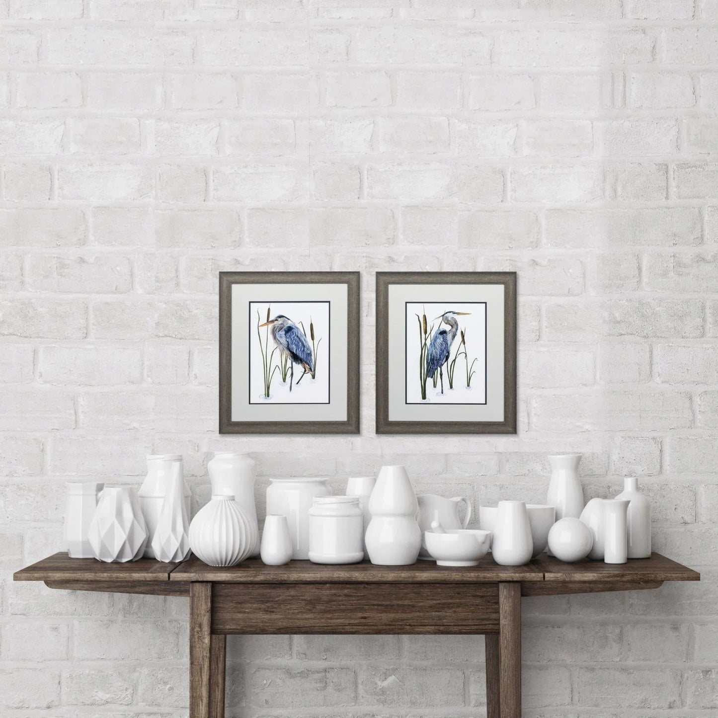 White ceramic vases on a wooden table against a white brick wall with framed artwork.